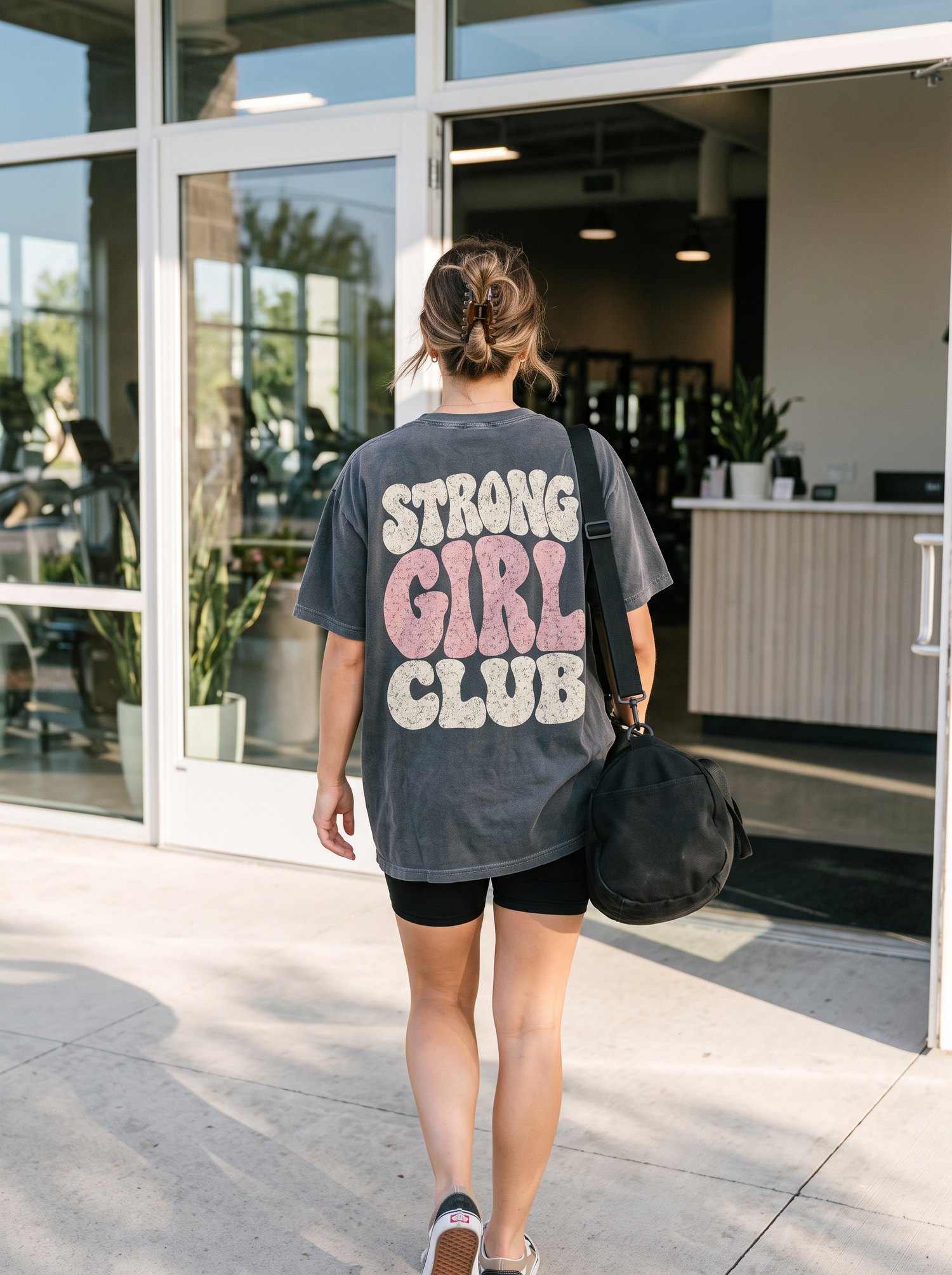 Woman walking into the gym with a black gym bag, wearing a Strong Girl Club oversized tee and biker shorts