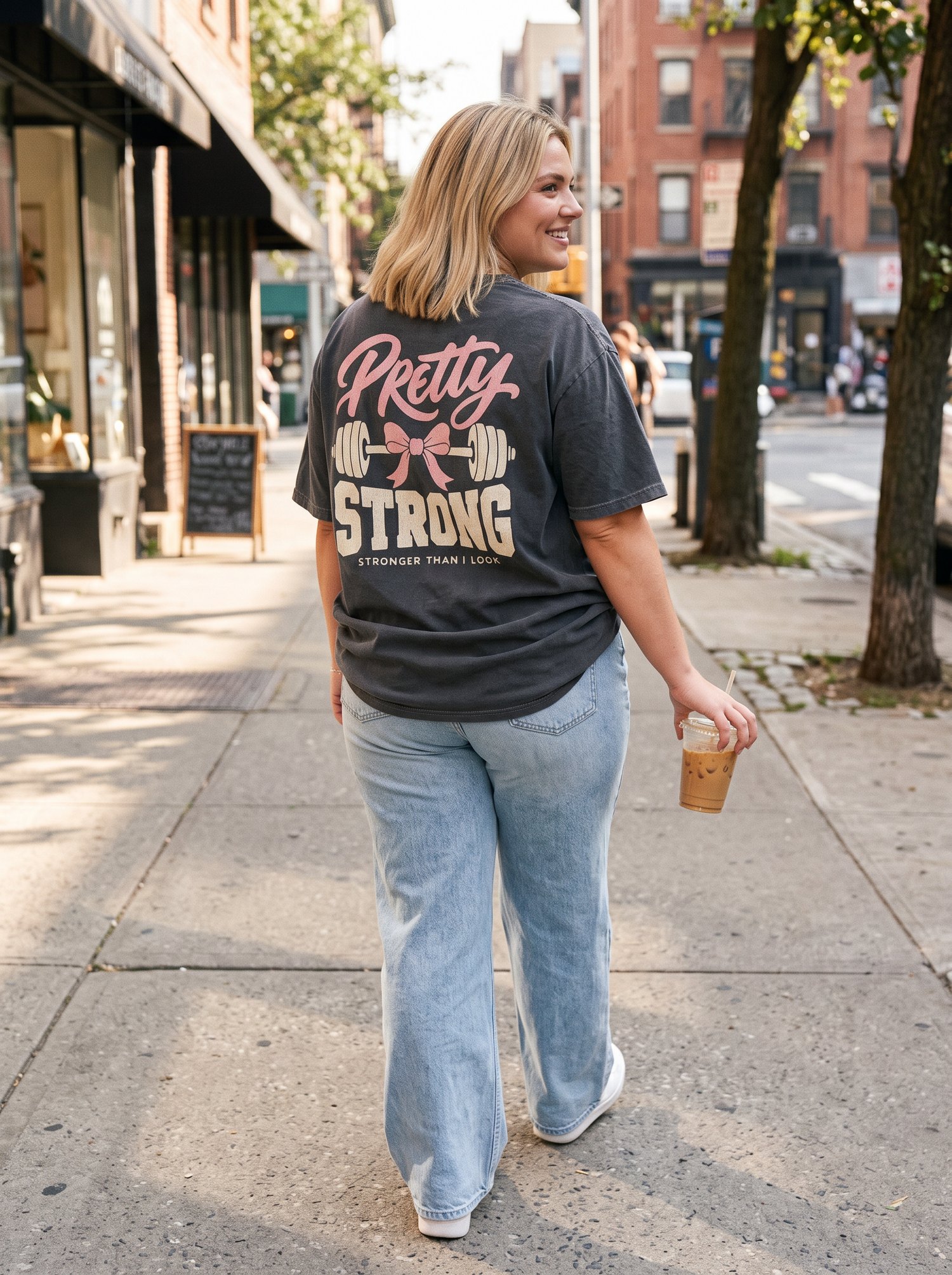 Woman in a Pretty Strong oversized tee with wide-leg jeans and an iced coffee