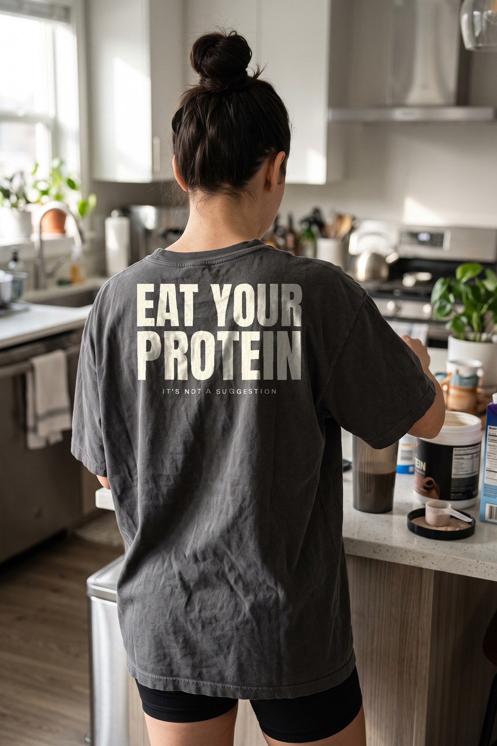 Woman making a protein shake in the kitchen wearing an Eat Your Protein oversized tee